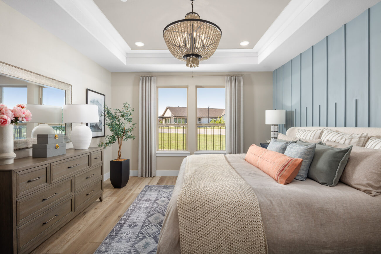 Elegant bedroom with tray ceiling, chandelier, blue accent wall, large windows, neutral decor, and cozy bedding with throw pillows