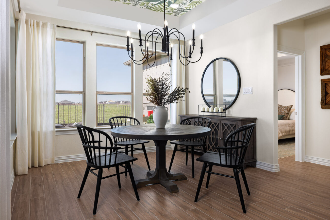 Staged dining area with round table, four black chairs, chandelier, large windows, and sideboard with round mirror