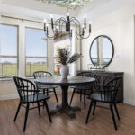 Staged dining area with round table, four black chairs, chandelier, large windows, and sideboard with round mirror
