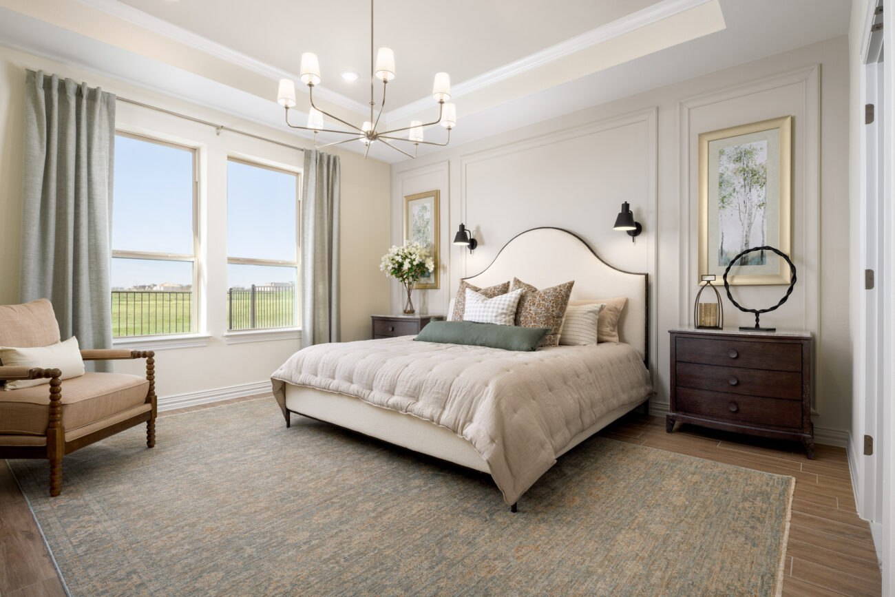 Staged primary bedroom with upholstered bed, large rug, modern chandelier, and two windows with curtains