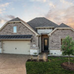 Brick single-story Ladera home with two-car garage, landscaped yard, and sunset sky in the background