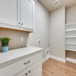 Bright utility room at Ladera Living with white cabinetry, built-in shelving, laundry hookups, and wood-style flooring