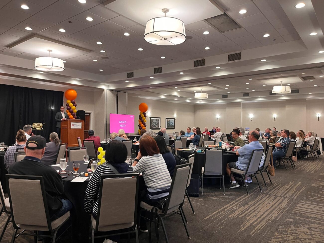 Several groups of people sit at tables watching a presentation in a large ballroom. A Ladera Living representative is presenting next to a large screen reading "What's Next"