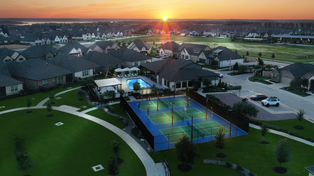 Aerial view over Ladera at Tavolo Park 55+ community in Fort Worth. Four pickleball courts cam be seen in the foreground, with an amenity center, pool, and dozens of homes in the background. The sun is setting in the distance.